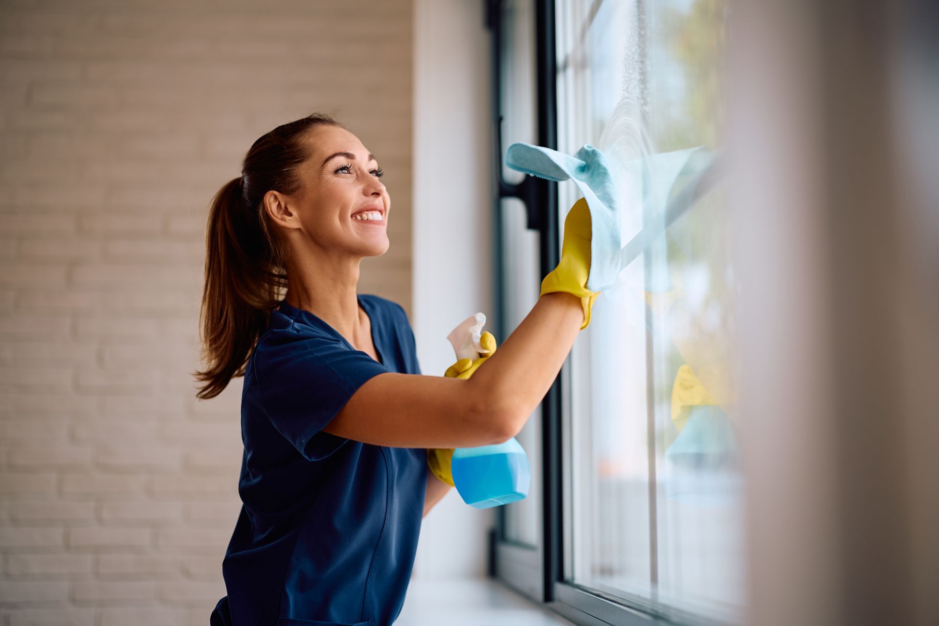 A smiling person in a blue uniform and yellow gloves cleaning a window with a spray bottle and cloth.