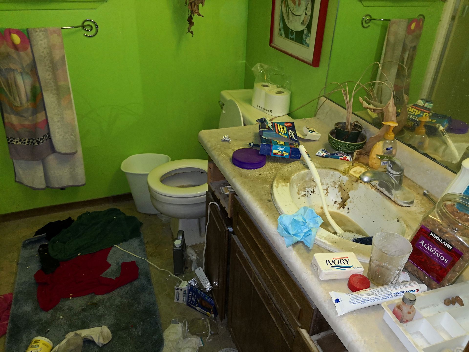 Cluttered bathroom with bright green walls, a messy sink vanity, and clothes scattered on the floor rug.