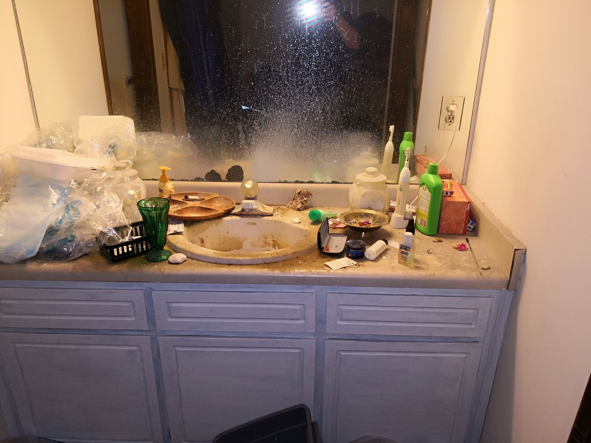 A bathroom vanity with a cluttered countertop, a stained sink, and a smudged mirror above grey cabinets.