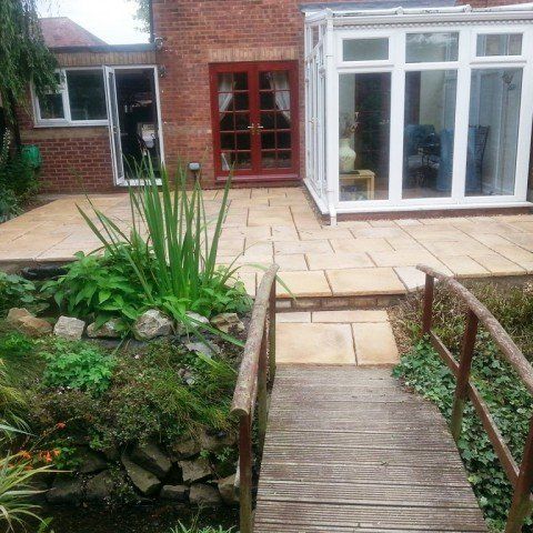A wooden bridge leads to a patio in front of a house.
