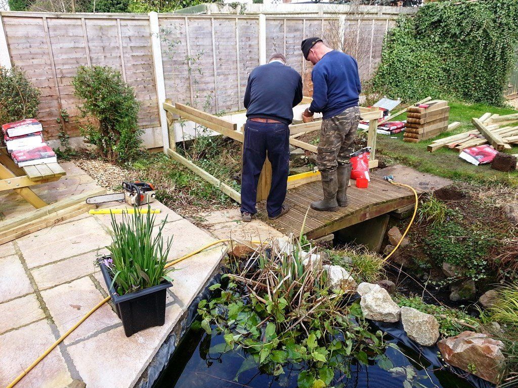 Two men are working on a wooden bridge over a pond.