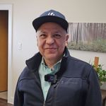 Man smiling indoors, wearing a navy cap and dark jacket with a small camera on his chest.