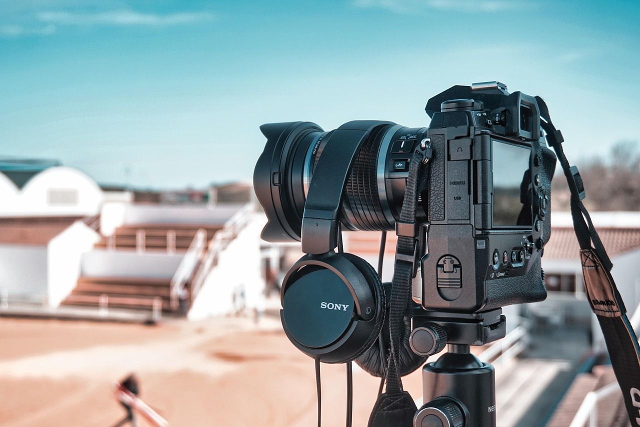 Camera on tripod with headphones, outdoors on a sunny day, focusing on a distant subject near white buildings.