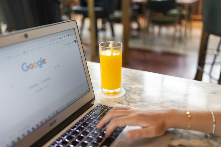Person typing on a laptop with Google homepage open; a glass of orange juice sits nearby.