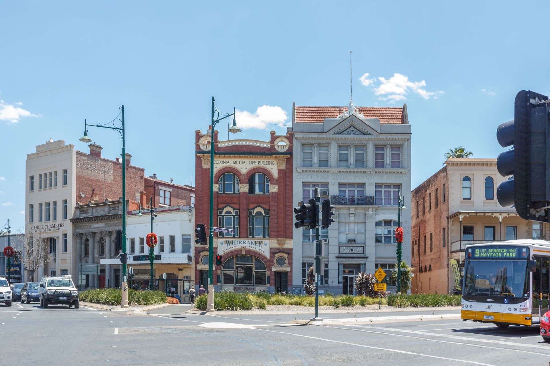 A sunlit city intersection featuring historic, multi-story buildings, traffic lights, and a public bus.