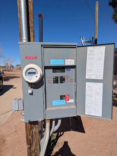 Exterior electrical panel on a pole, door open, with circuit breakers and meter, on a sunny day.