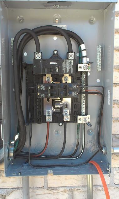 Gray electrical panel mounted on a brick wall, showing wires and circuit breakers.