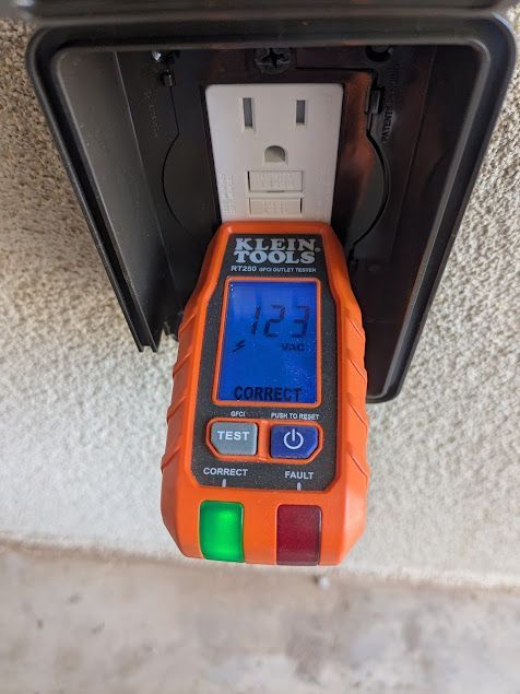 A Klein Tools outlet tester plugged into an outdoor outlet, reading 123 volts. The tester shows a green light.