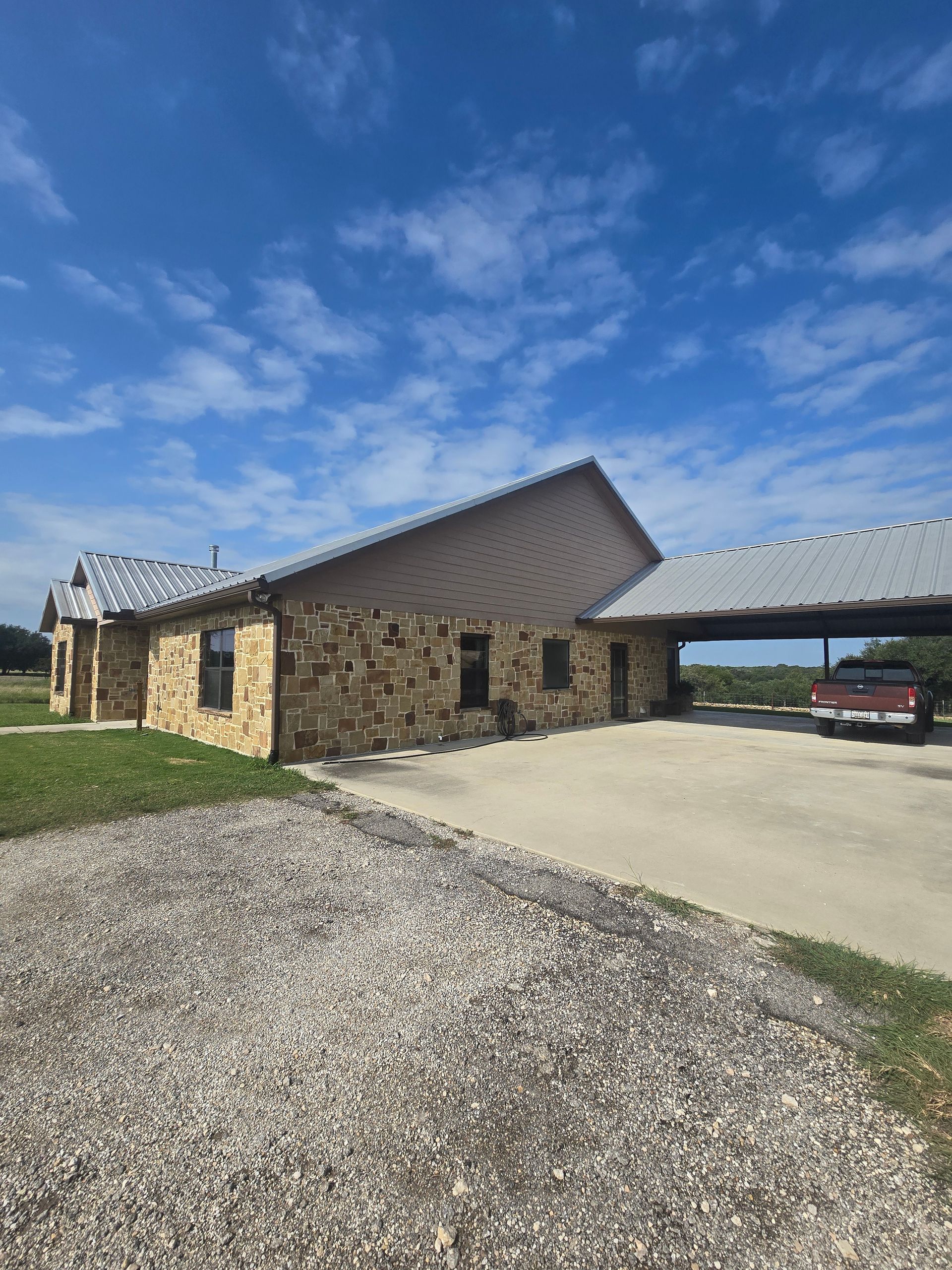 Stone building with carport, driveway, blue sky, and a truck parked under the carport.