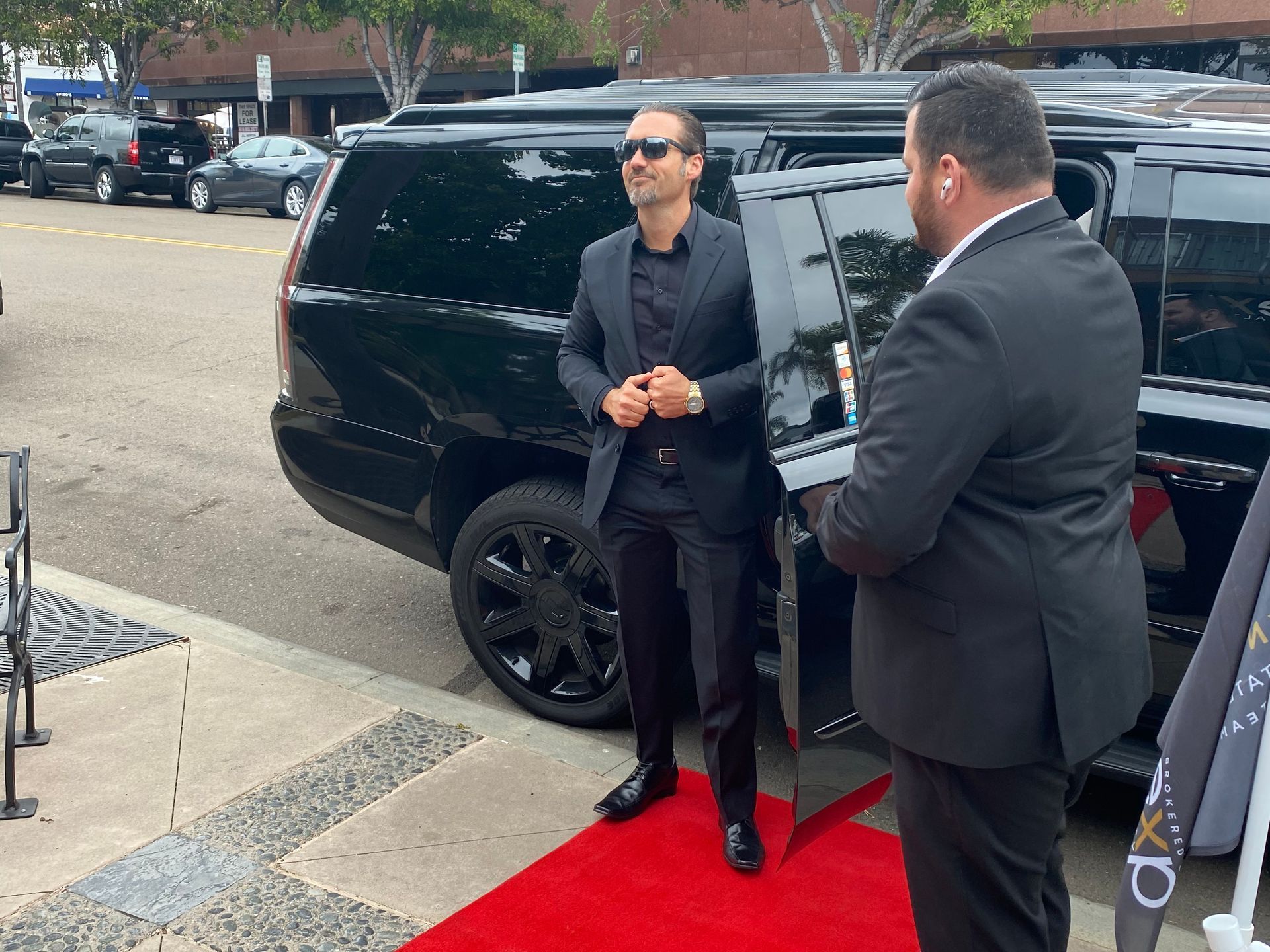 two men in suits are standing next to a black suv on a red carpet .