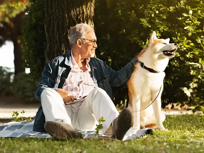 Un anciano está sentado sobre una manta en el césped con su perro.