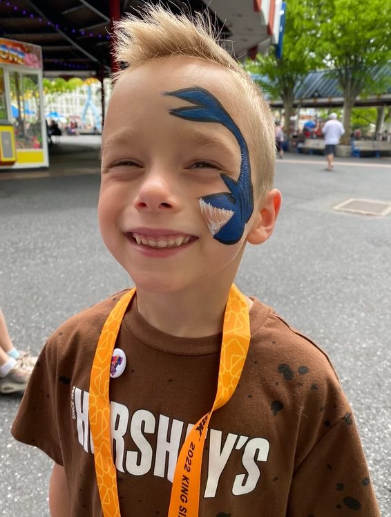 A smiling child with a blue shark painted on their face, wearing a brown Hershey's t-shirt at an outdoor park.