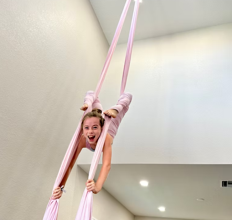 A person hanging upside down from two suspended pink fabric silks, smiling broadly while posing in a room.