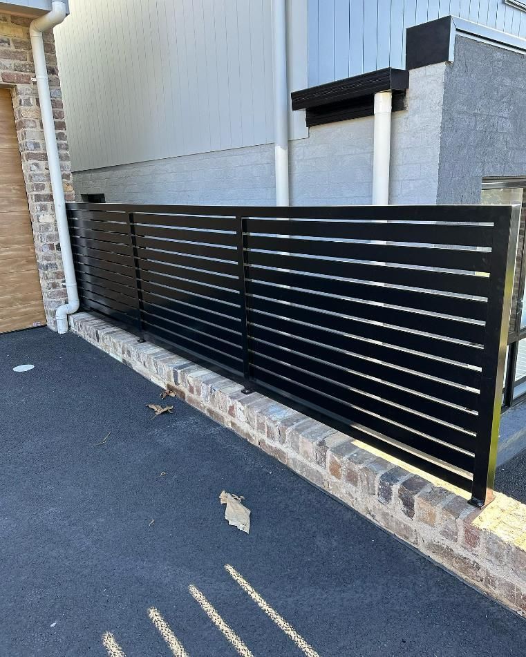 A Black Fence Is Sitting Next to A Brick Wall — Optimum Aluminium Fabrication in Barrack Heights, NSW