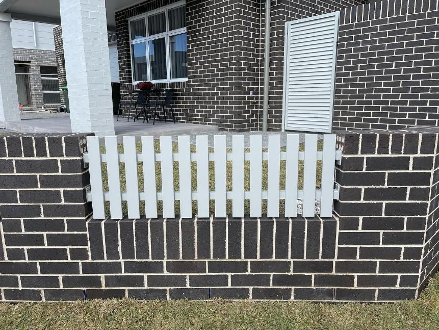 A Brick Wall with A White Picket Fence in Front of A House — Optimum Aluminium Fabrication in Barrack Heights, NSW