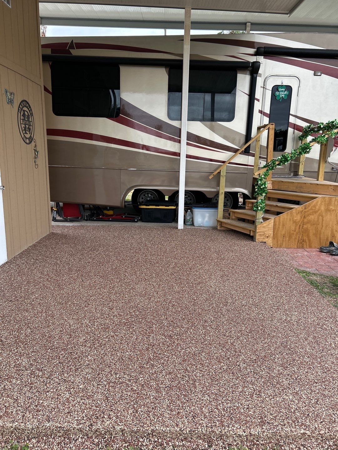 RV parked under a carport with a gravel patio. Steps lead to the RV door, garland adorns the railing.