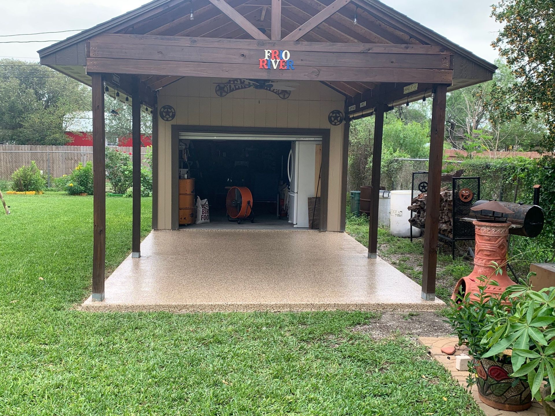 Garage with open door, carport. Concrete floor. Surrounded by grass, fence, plants.