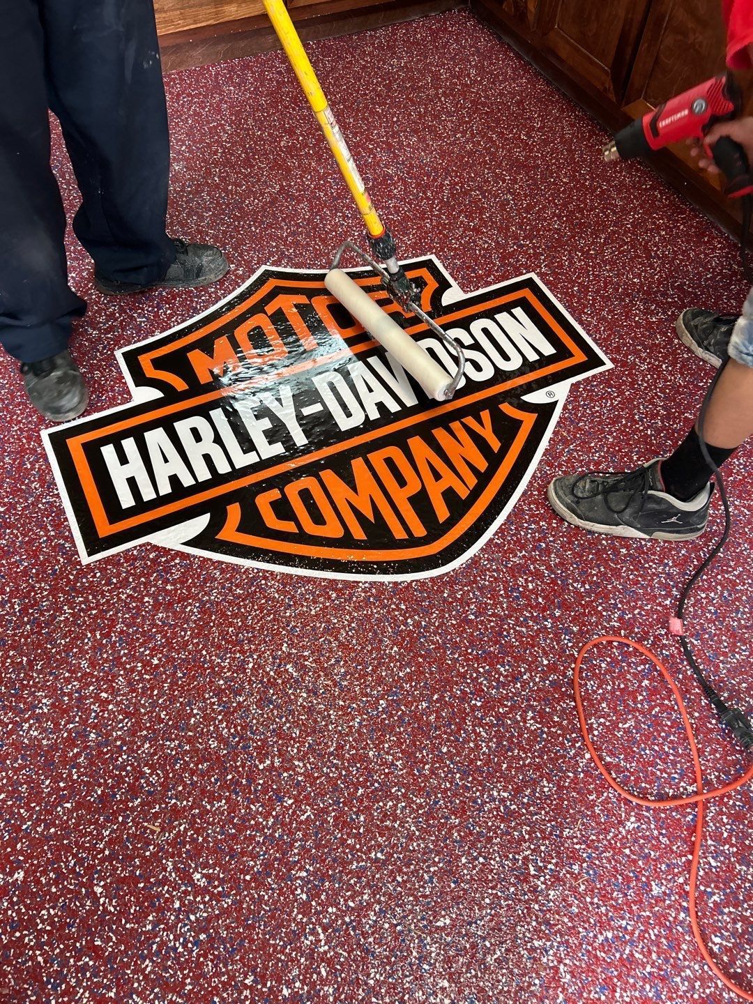 Two people applying epoxy floor finish with a Harley-Davidson logo inlay. Red speckled floor.