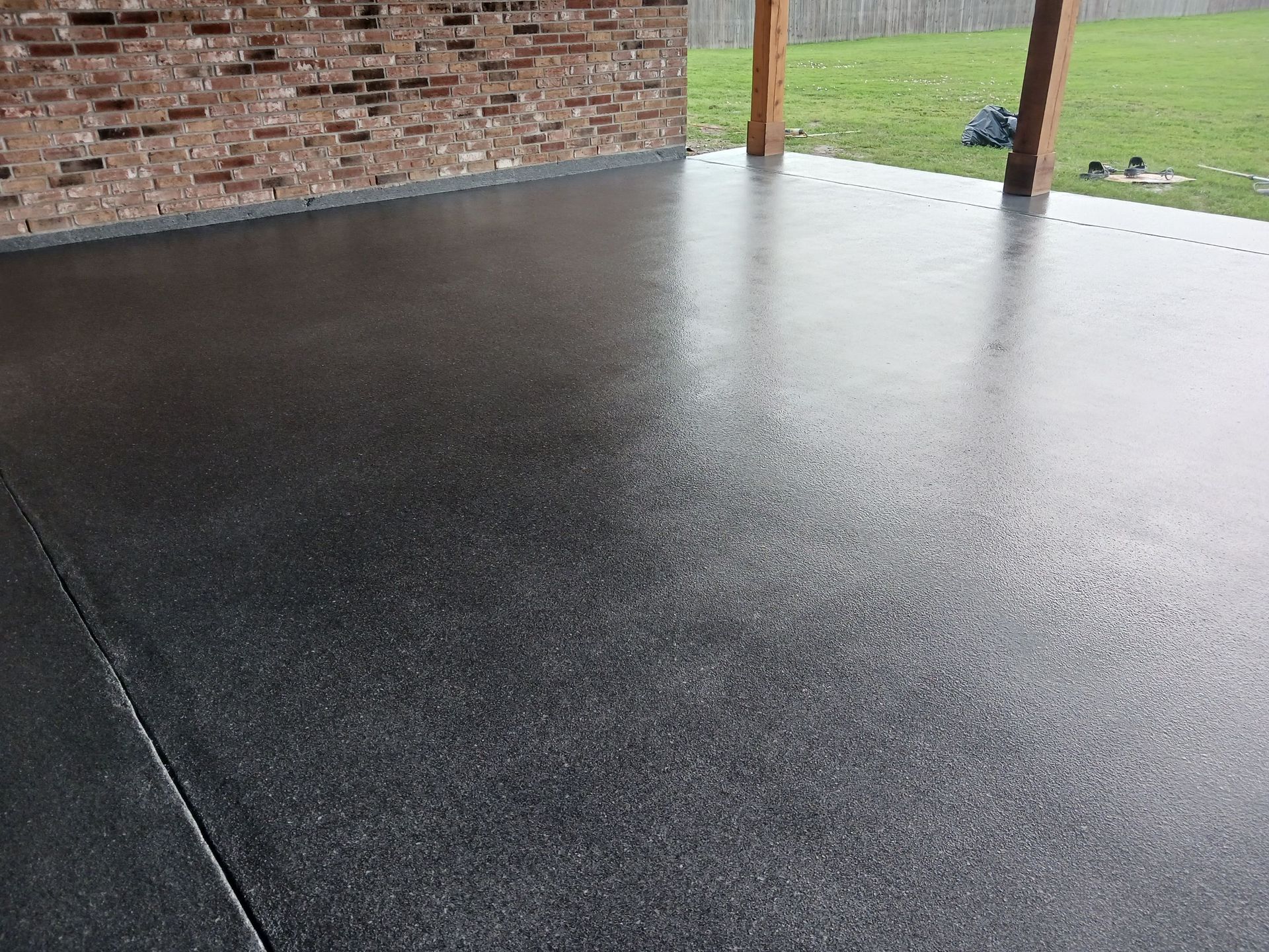 Wet, dark gray concrete patio with brick wall and wooden posts, overlooking grass.