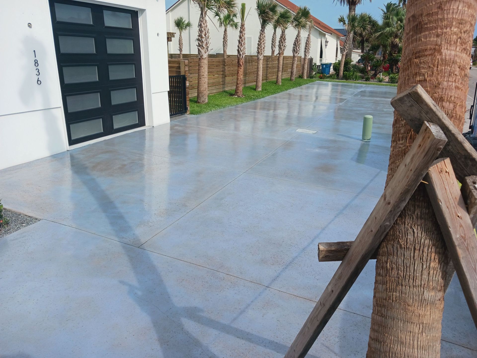 A gray concrete driveway leads to a modern home with a black garage door, palm trees line the street.