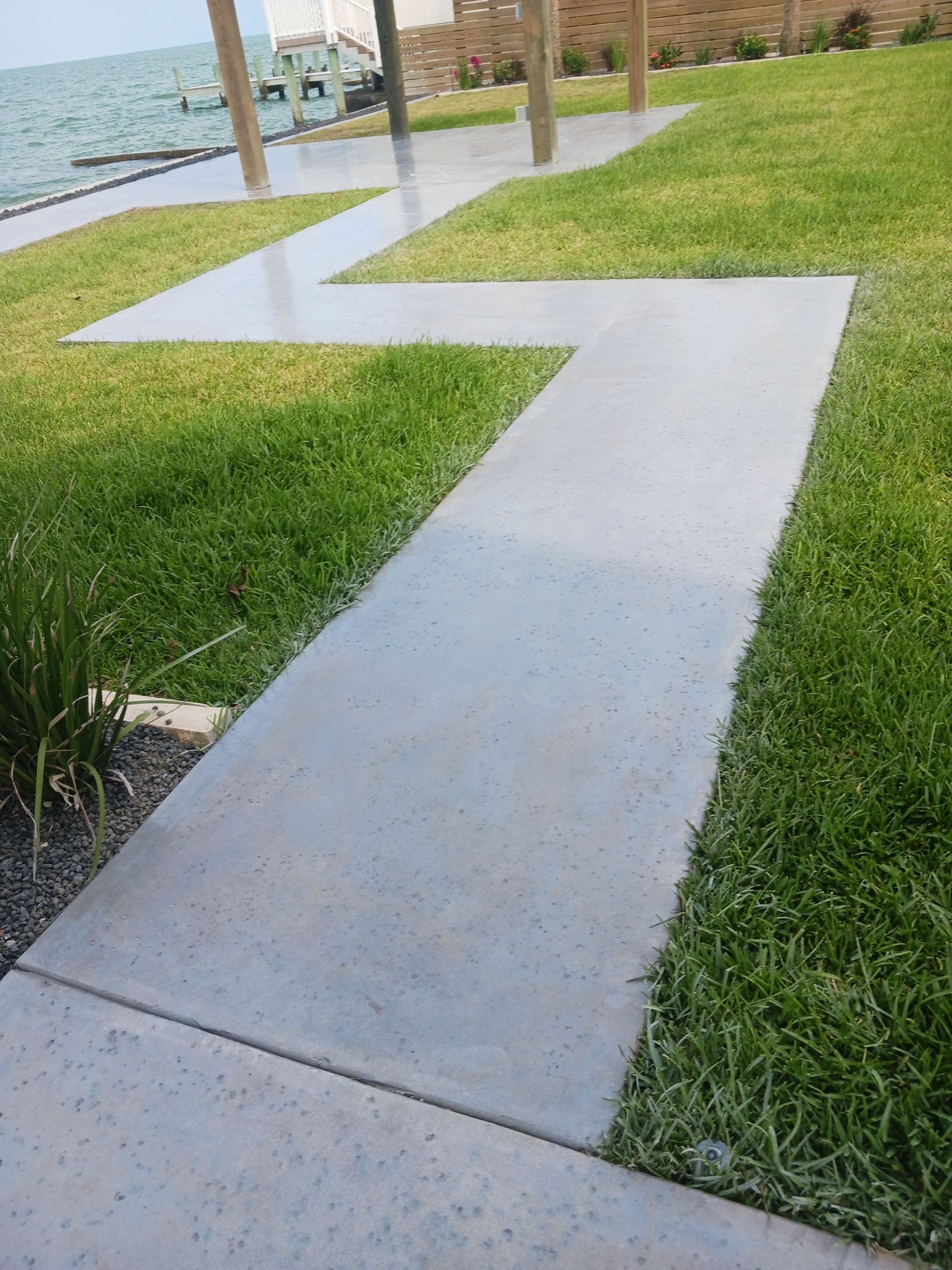 A zigzag concrete walkway through green grass, leading to an outdoor structure, next to water.