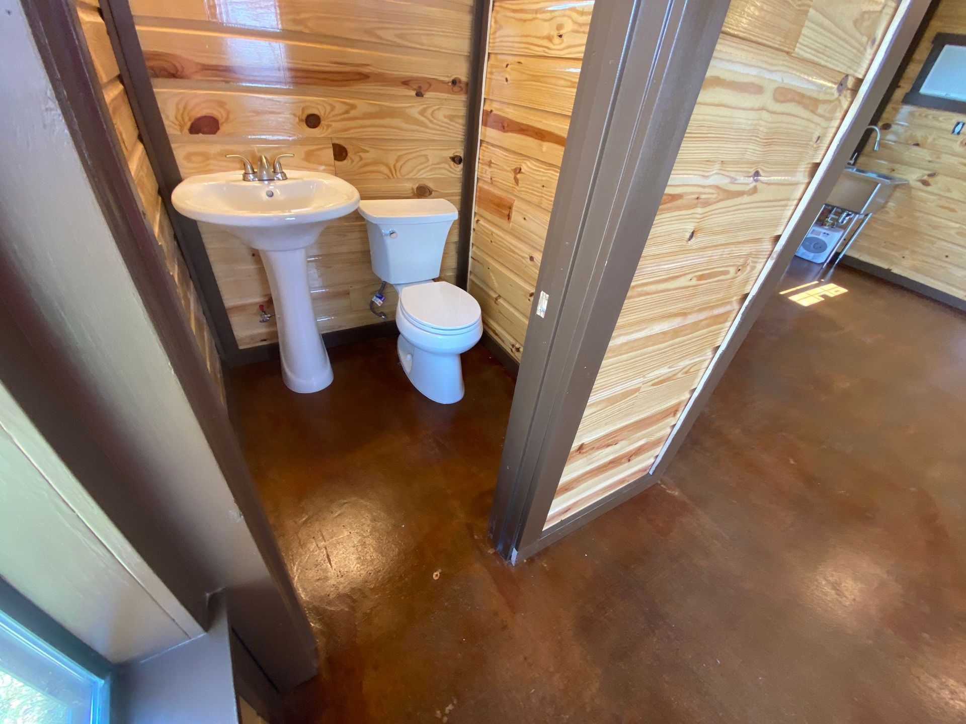 Small bathroom with a pedestal sink and toilet, brown flooring, wooden walls.