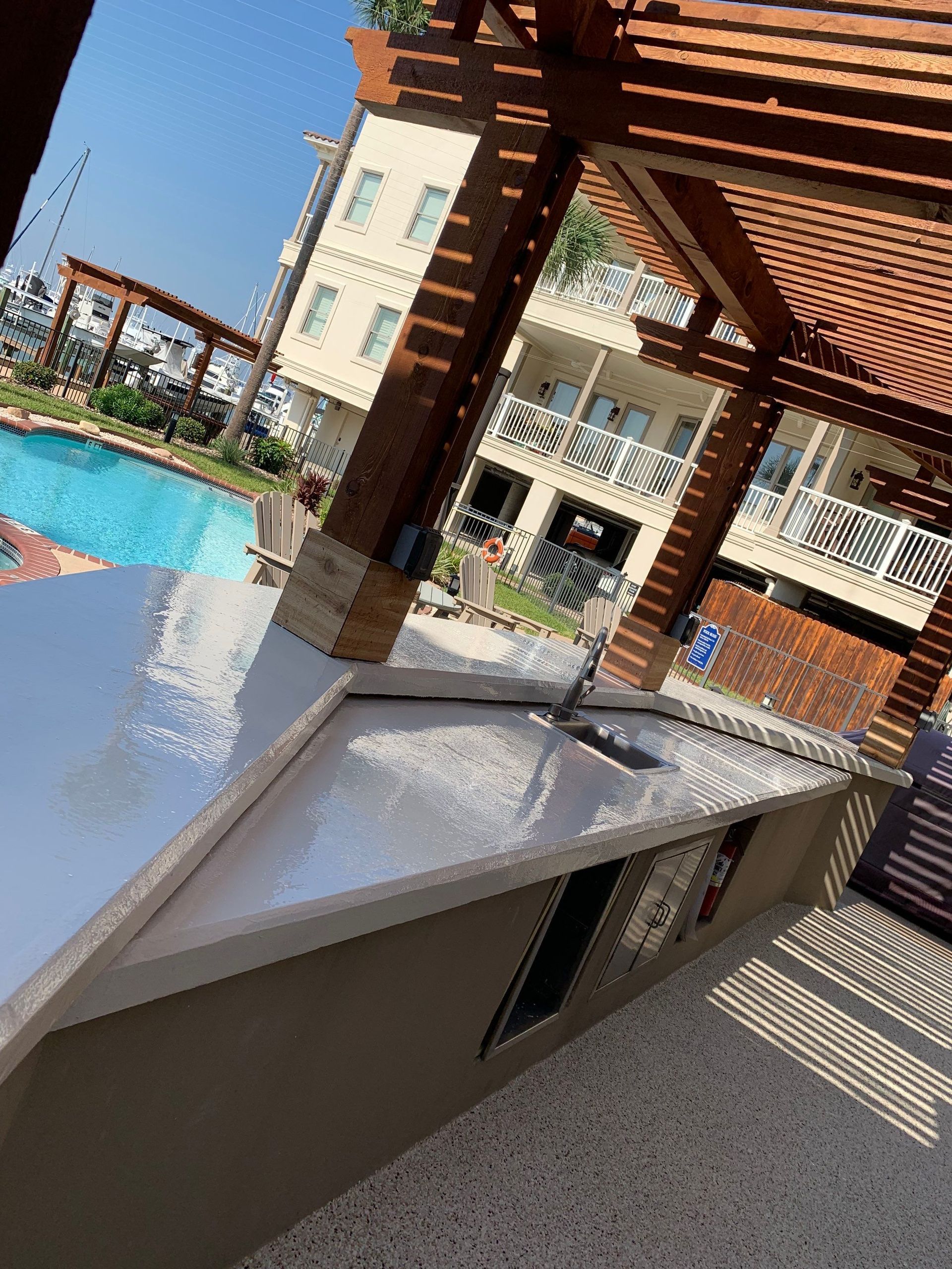 Outdoor kitchen next to a pool. Brown pergola, buildings in background, blue pool, sunny day.
