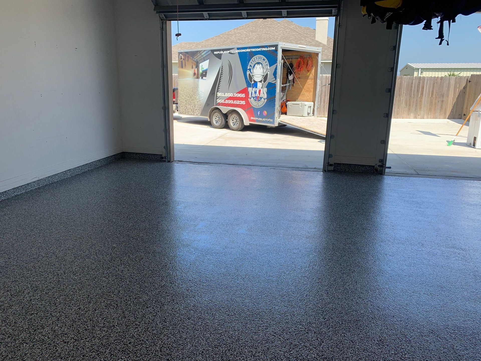 Garage with a glossy, speckled floor, open to a sunny exterior with a trailer.