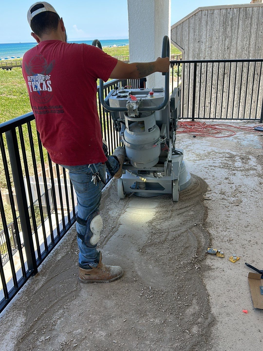 Man grinding concrete patio. Red shirt, blue jeans, knee brace. Outdoors, seaside background.