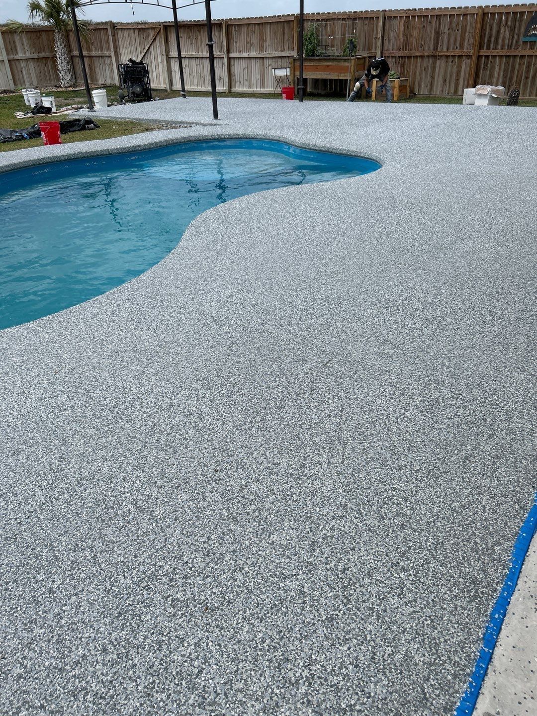 Poolside with a pebble-textured concrete surface. The pool has blue water. A wooden fence and yard with greenery.