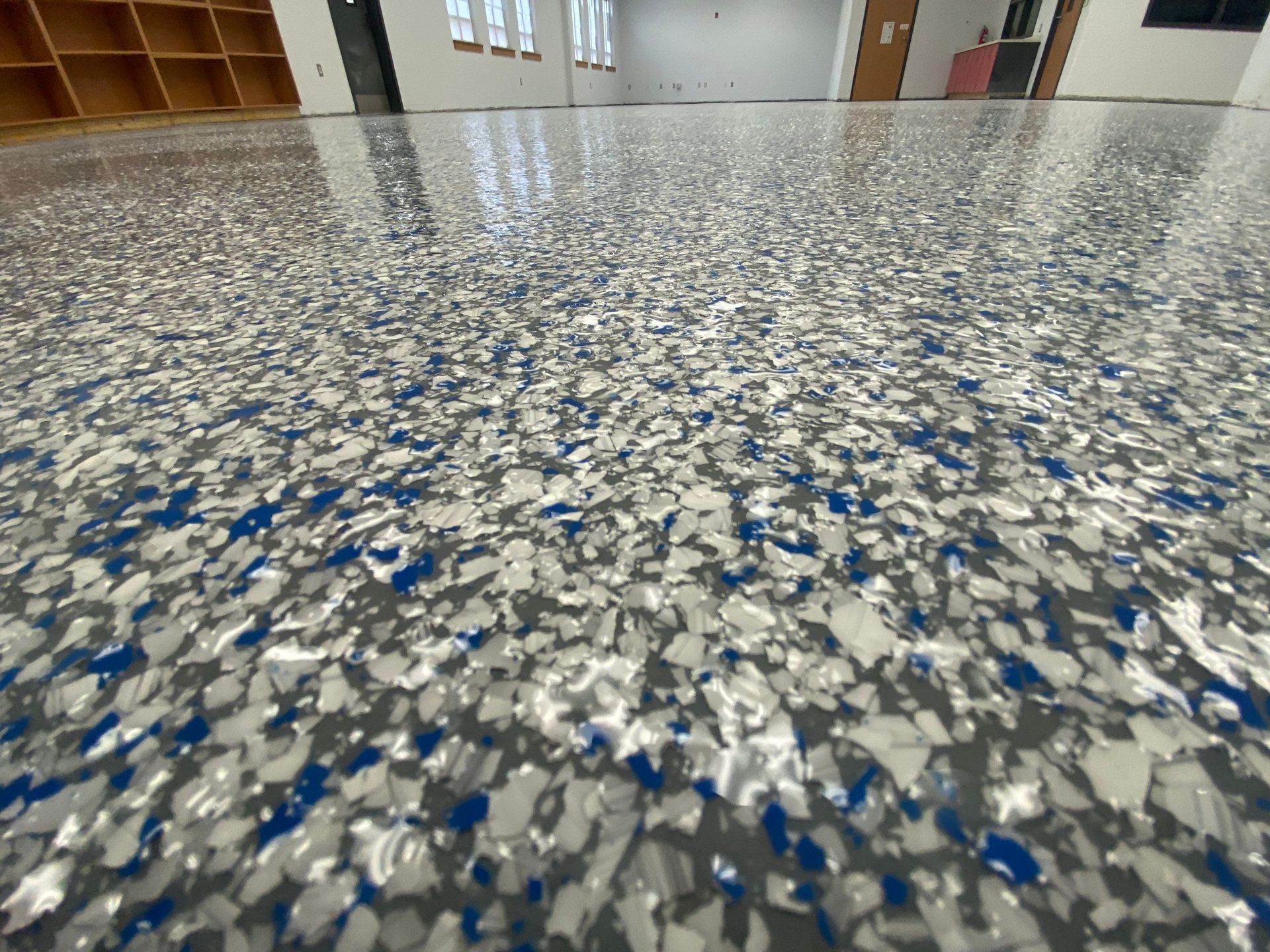 Close-up of a glossy floor with blue, gray, and white chips, inside a room with shelving and windows.