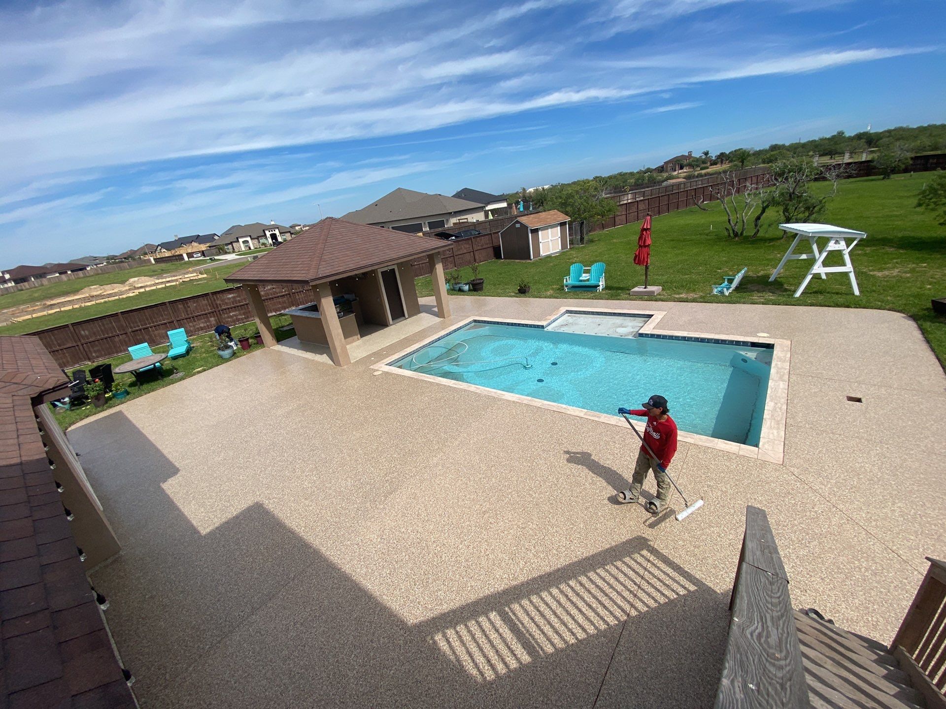 Person cleaning a pool on a sunny day. Brown patio, blue sky, and gazebo in the backyard.
