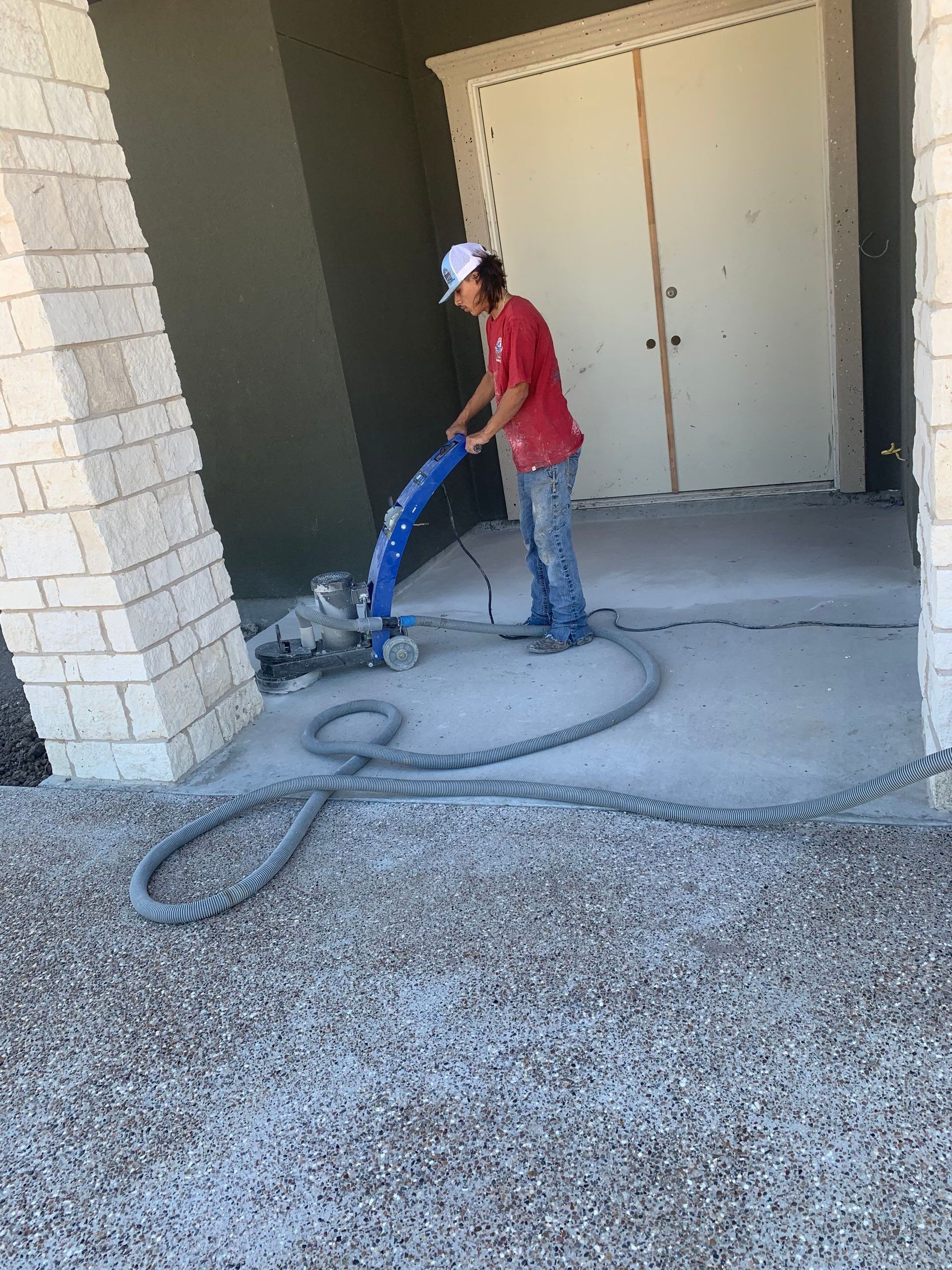 Man using a blue nozzle to work on a concrete surface. He is wearing a red shirt and jeans in a stone and concrete setting.