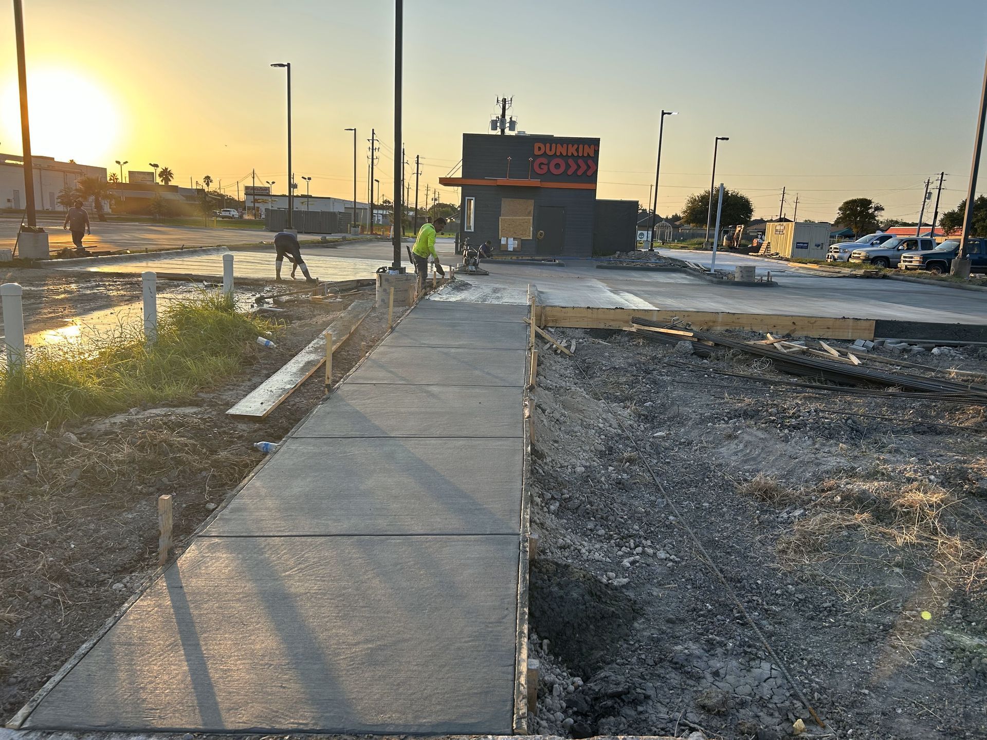 Construction site: Workers pouring concrete sidewalk near a building with orange and black signage during a sunrise.