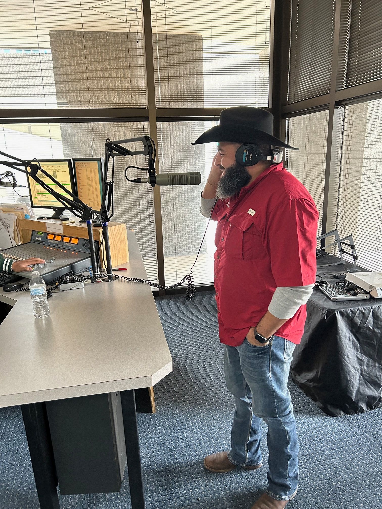 Man in red shirt, cowboy hat in radio booth, speaking into a microphone.