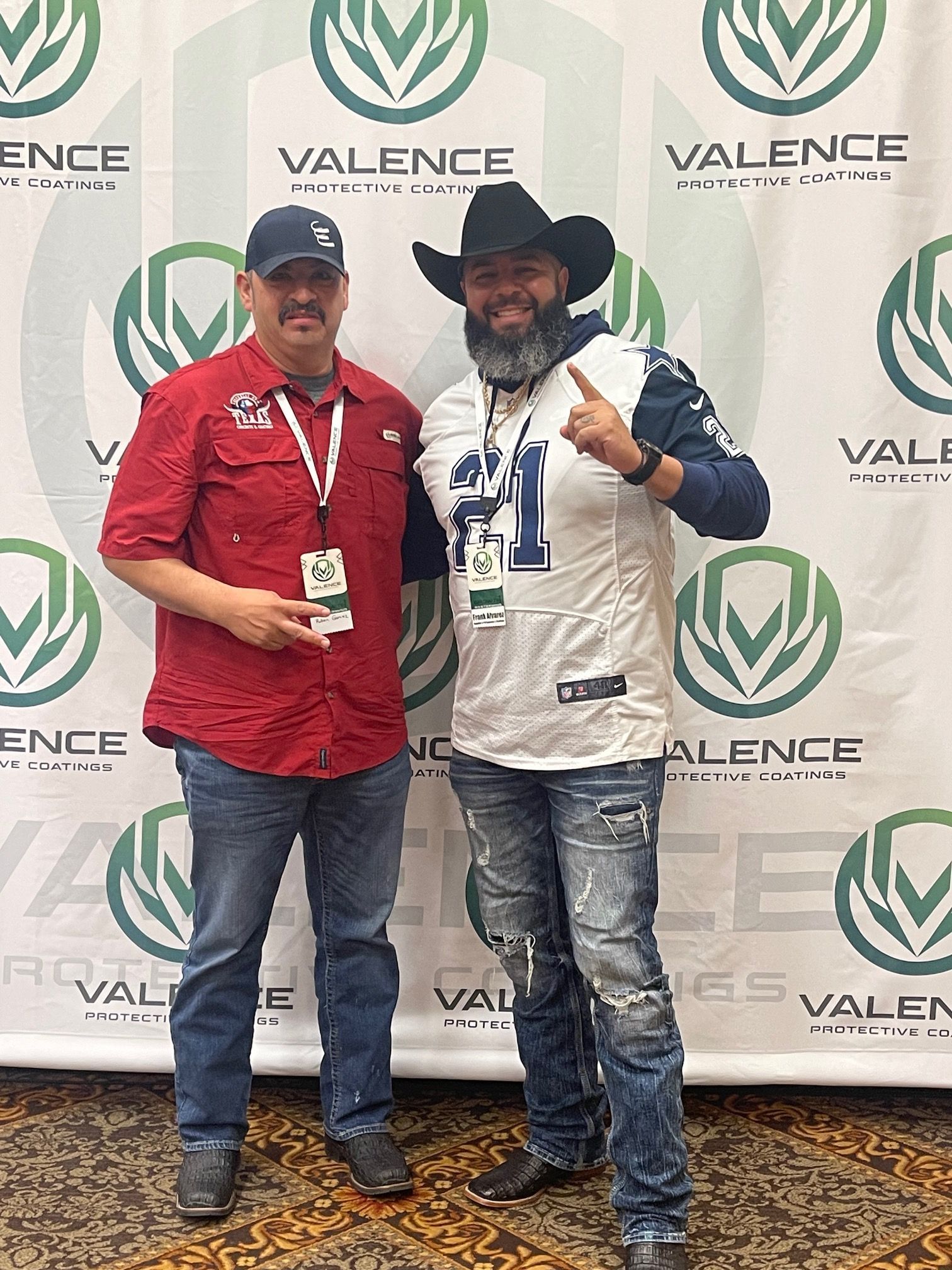 Two men posing for a photo in front of a VALENCE banner. One in a cowboy hat and jersey, the other in a red shirt.