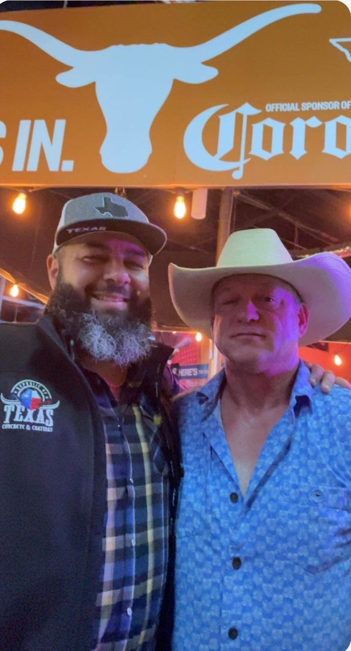  One wears a cowboy hat, the other a cap. Both smile in front of a sign with the Texas Longhorns logo.