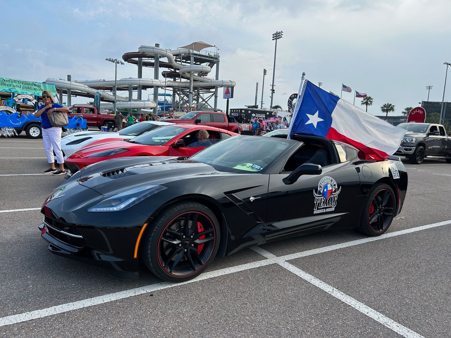 Black Corvette with Texas flag at car show, red sports car in background.