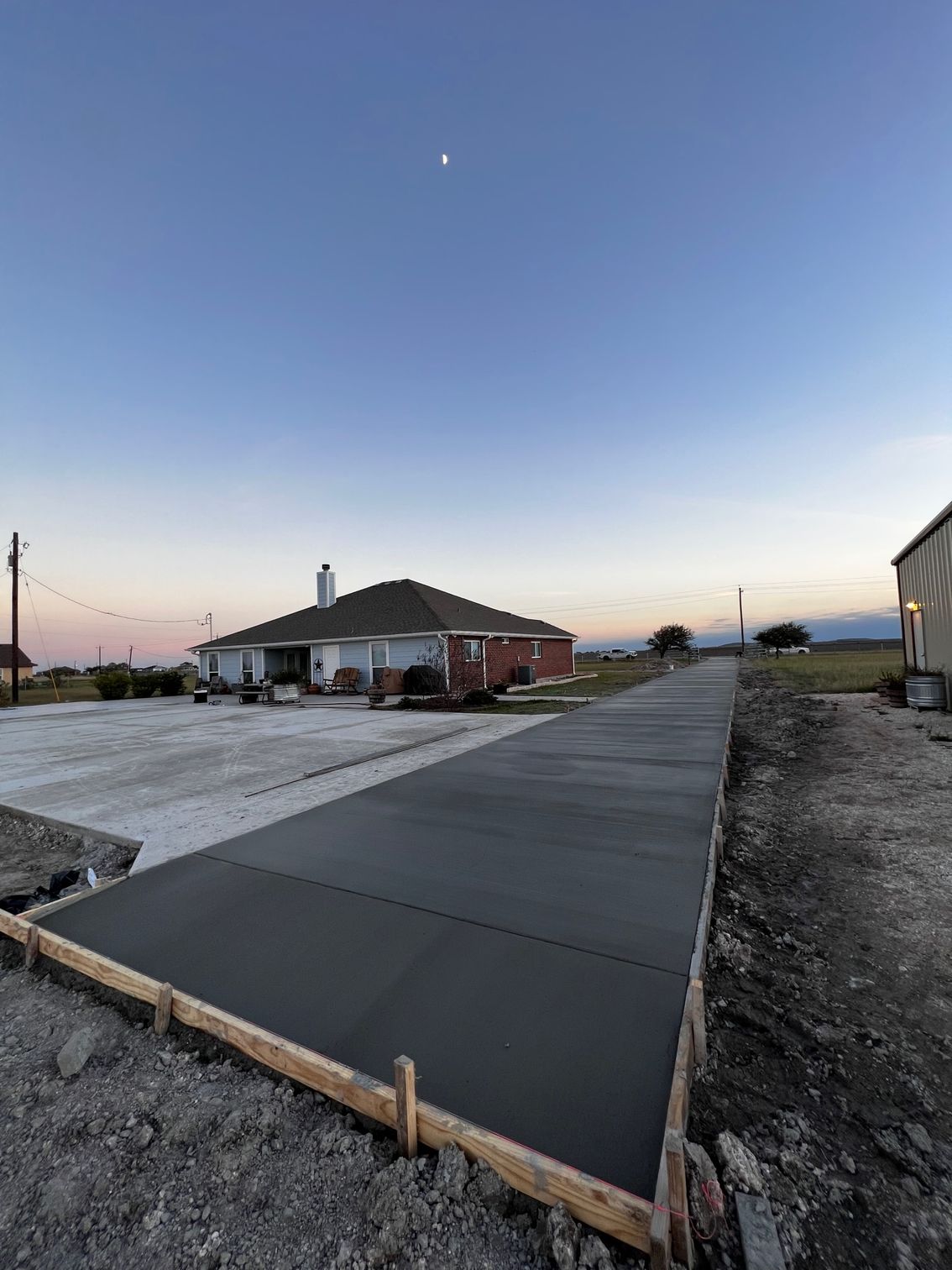 Newly poured concrete driveway leads to a brick house at dusk. Wooden forms line the edges, a half-moon.