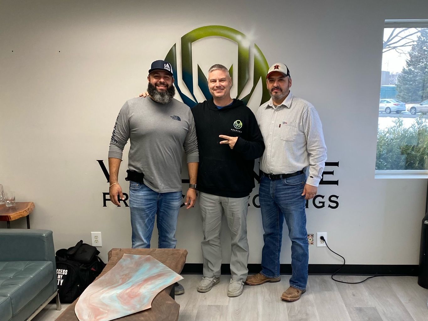 Three men stand in front of a business logo, smiling. One is making a peace sign. Interior shot.