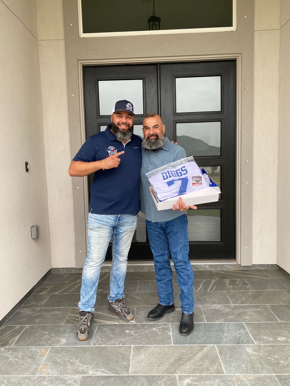 Two men smiling in front of a dark door. One points, the other holds a package labeled 
