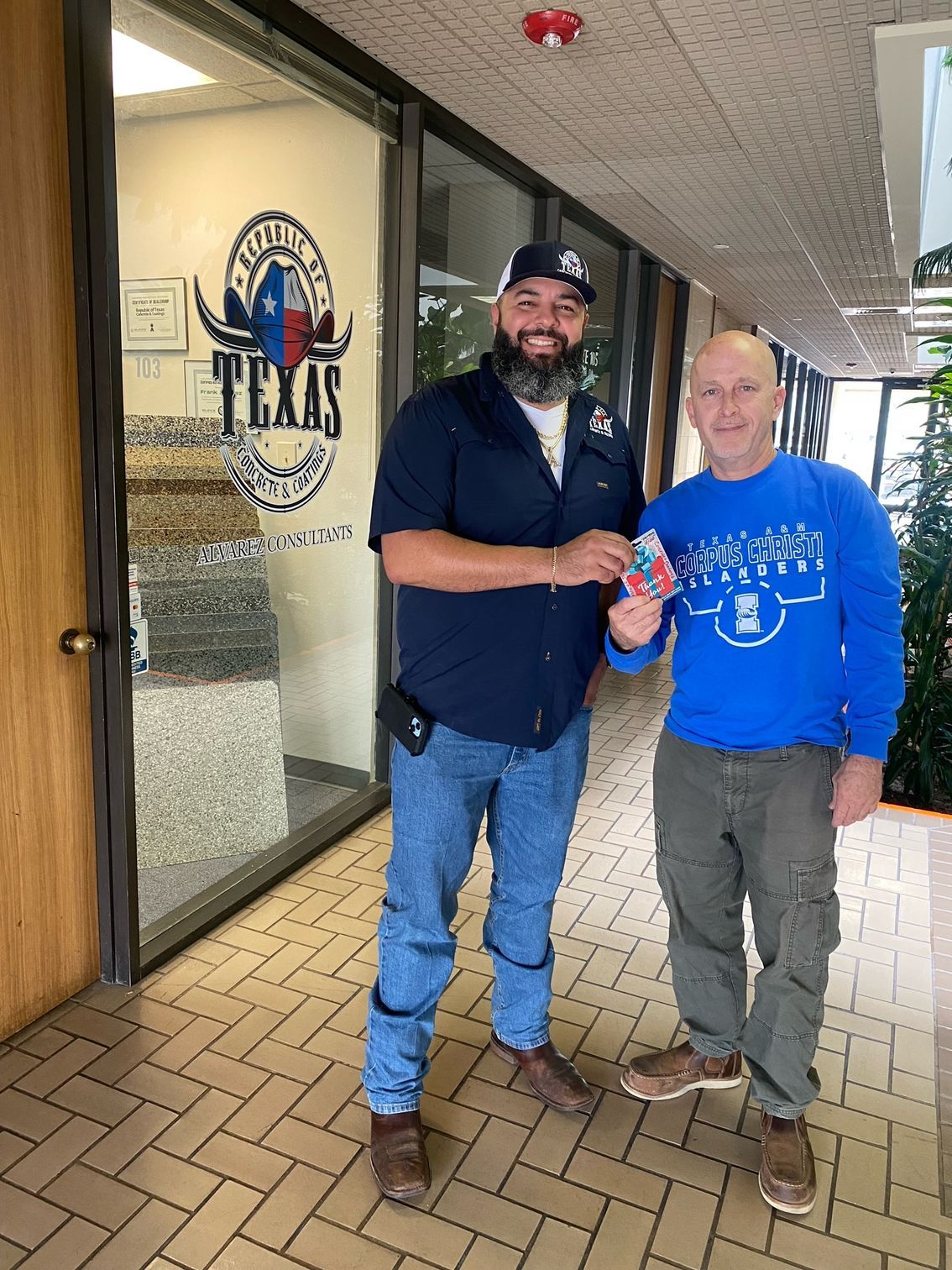 Two men in front of a Texas business: one holding a card, the other smiling.