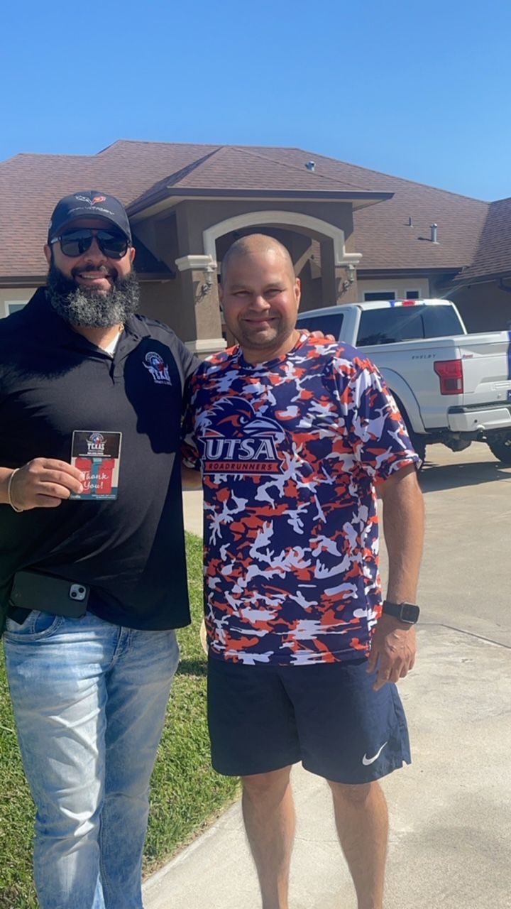 Two men smiling, posing in front of a house. One holds a UTSA hat and a cup, other wears a UTSA shirt.
