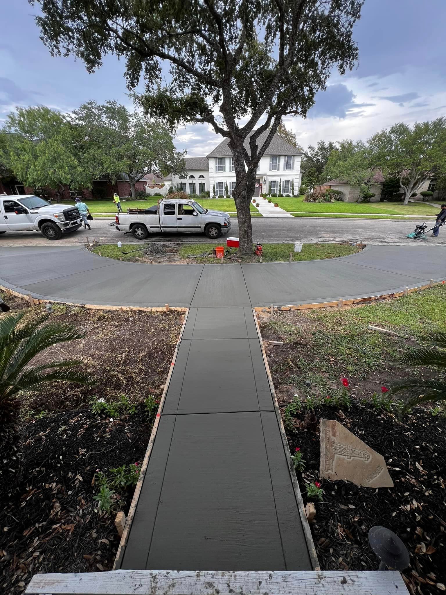 New concrete sidewalk and driveway leading to a two-story house. Trucks and workers visible in the background.