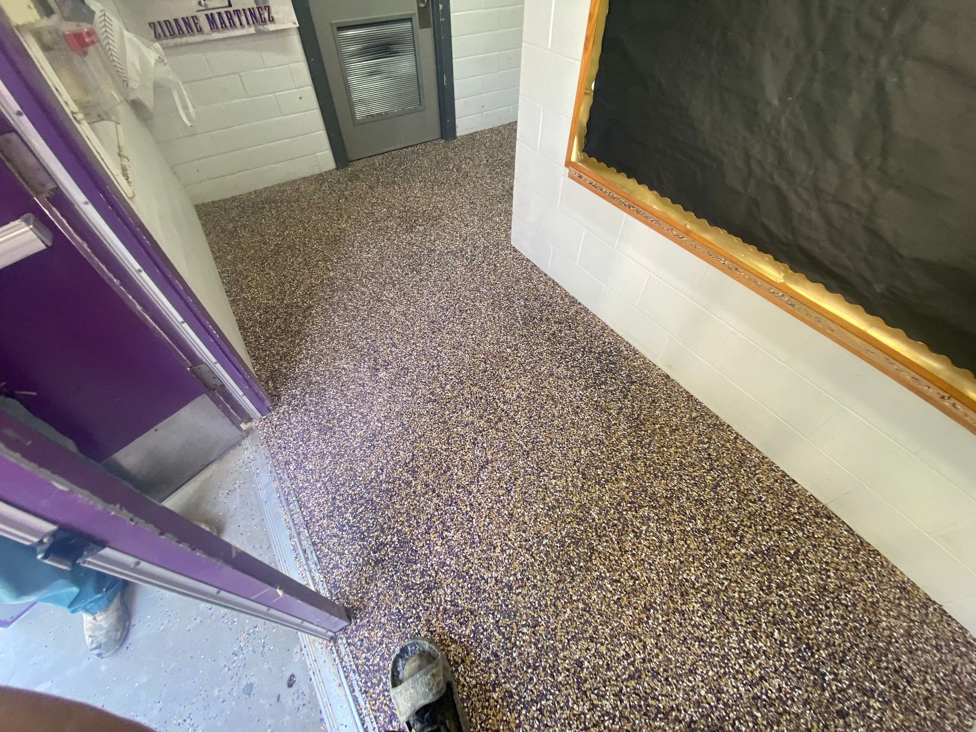 A hallway with a pebble-textured floor. Walls are white, purple door frame on the left, door and artwork on the right.