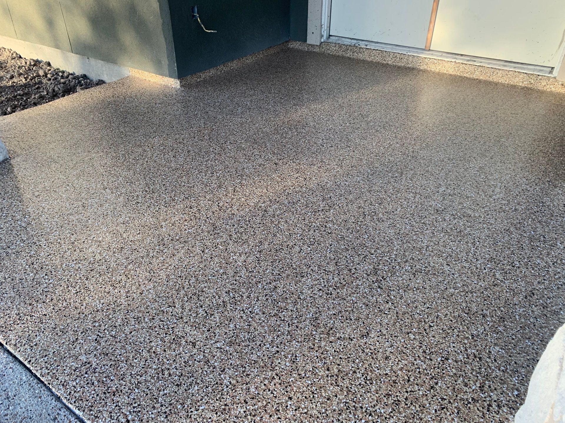 Epoxy-coated concrete patio with tan, brown, and gray chips, next to a light-colored door and dark wall.