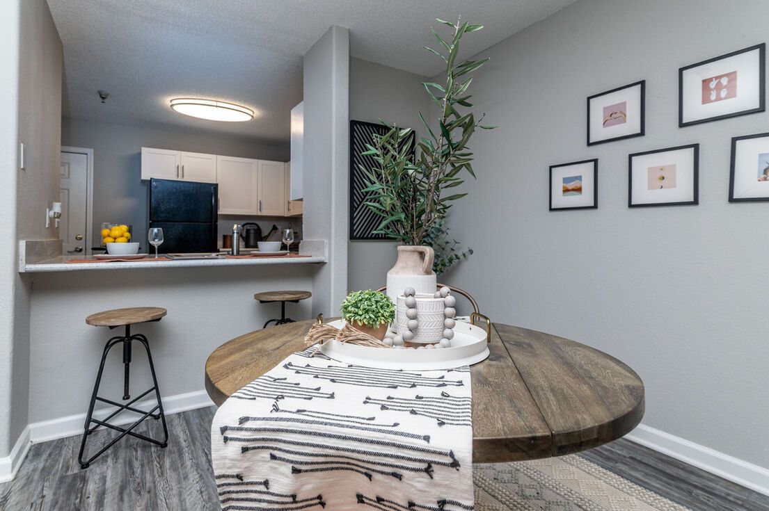 Dining area with round wooden table, stools, and kitchen in background. Gray walls, decor.