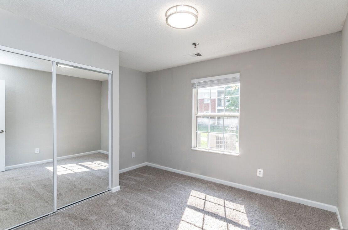 Empty gray bedroom with mirrored closet doors and a small window.