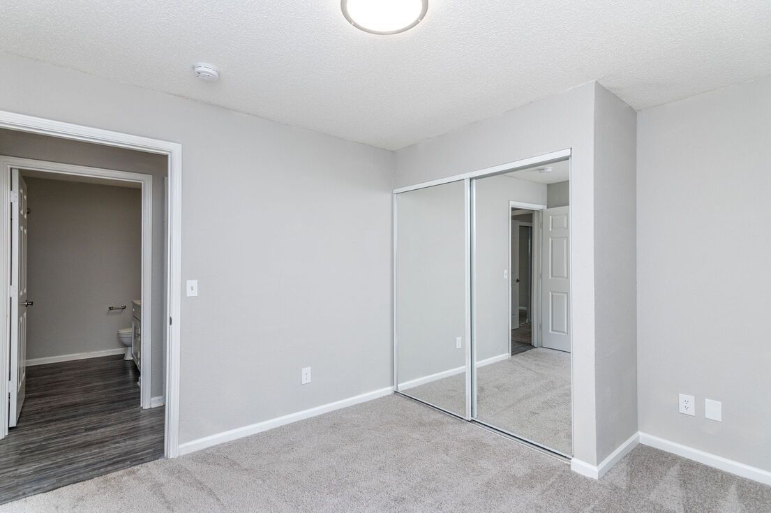 Empty bedroom with gray walls, carpet, mirrored closet doors, and a doorway to a bathroom.
