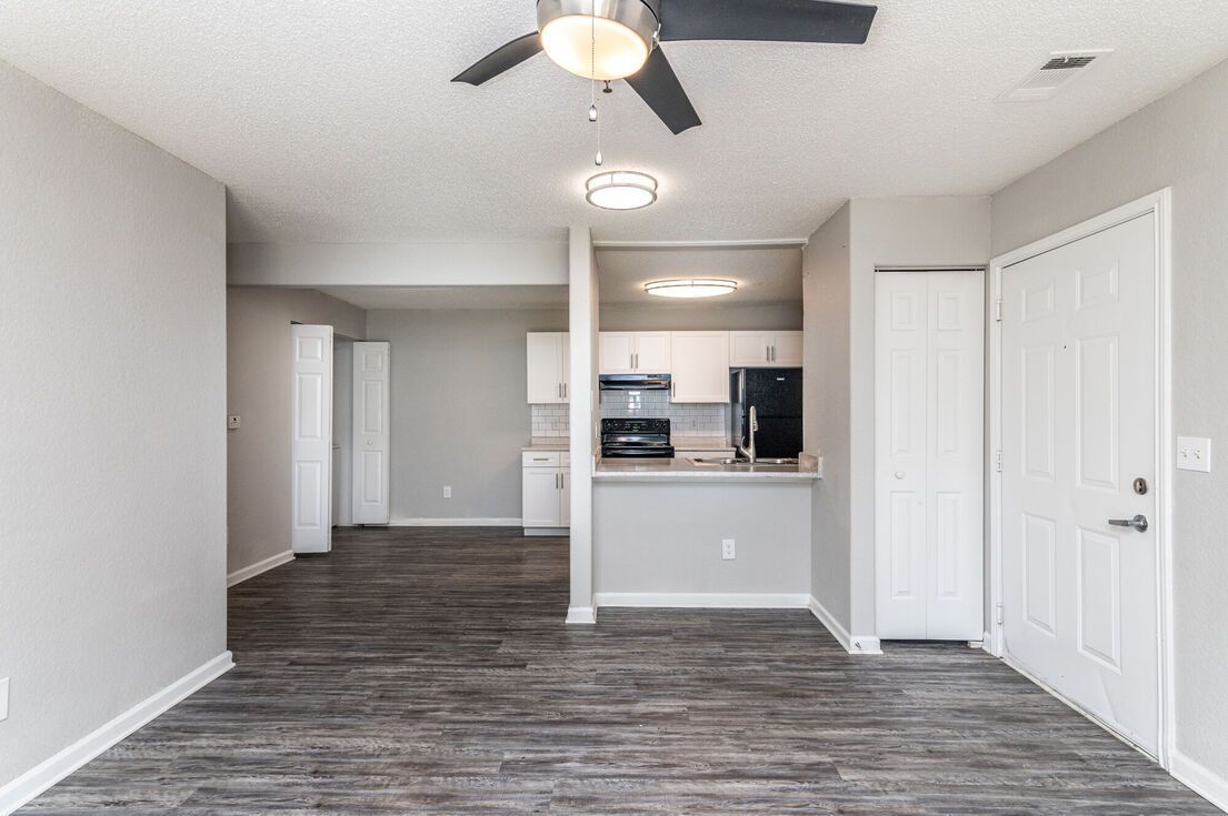 Living room with kitchen in the background; gray walls, dark flooring, and white cabinetry.